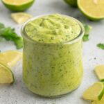 Jar of a creamy green dressing surrounded by limes and cilantro.