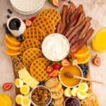 Breakfast and brunch charcuterie board with waffles, bacon, sausage, hard-boiled eggs, syrup, fruit, honey, toast, butter, and yogurt.