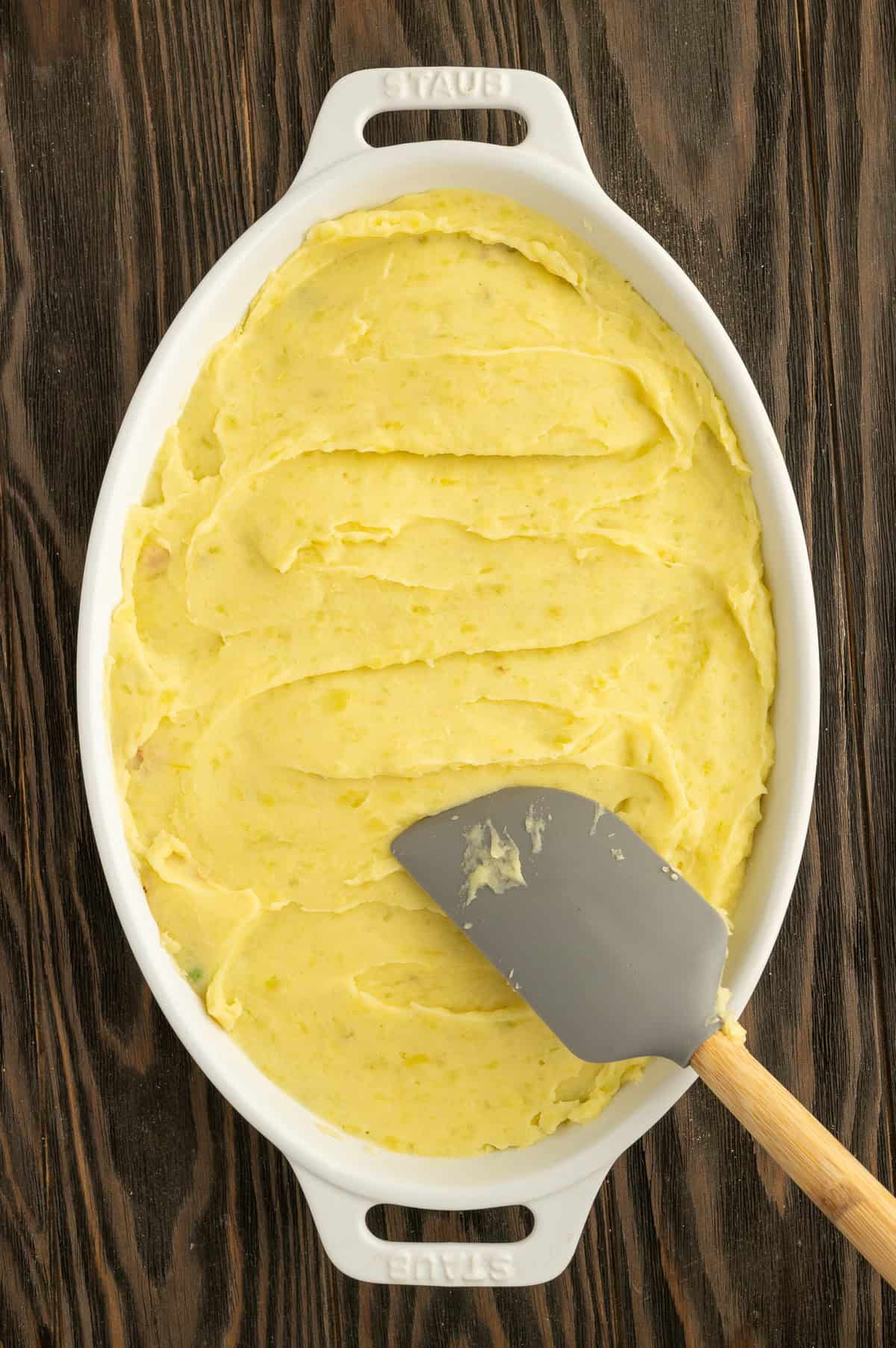 Spreading mashed potatoes over meat mixture in white casserole dish.
