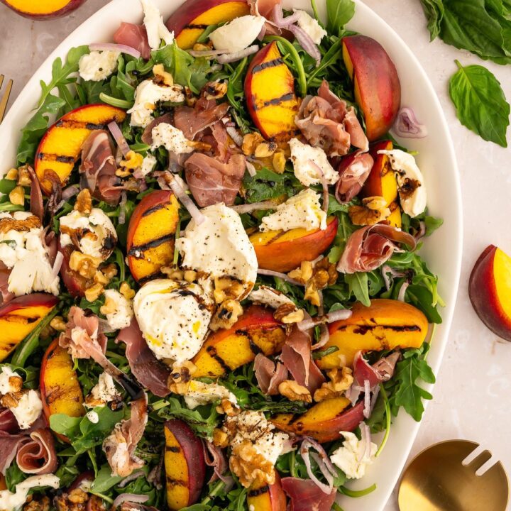 A salad with grilled peaches, burrata, prosciutto and balsamic dressing on a white platter.