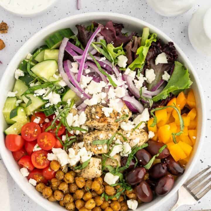 Bowl of Mediterranean ingredients like lettuce, red onion, feta, bell pepper, Greek chicane, tomato, olives and chickpeas that shows how to assemble a meal prep lunch.