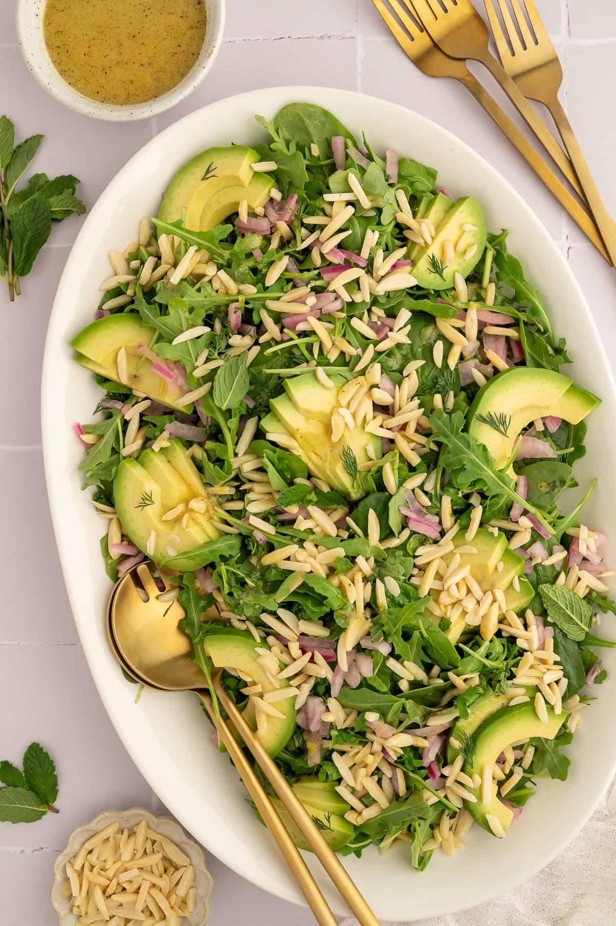 Salad on a white platter with arugula, spinach, slivered almonds, dill, mint, and avocado.