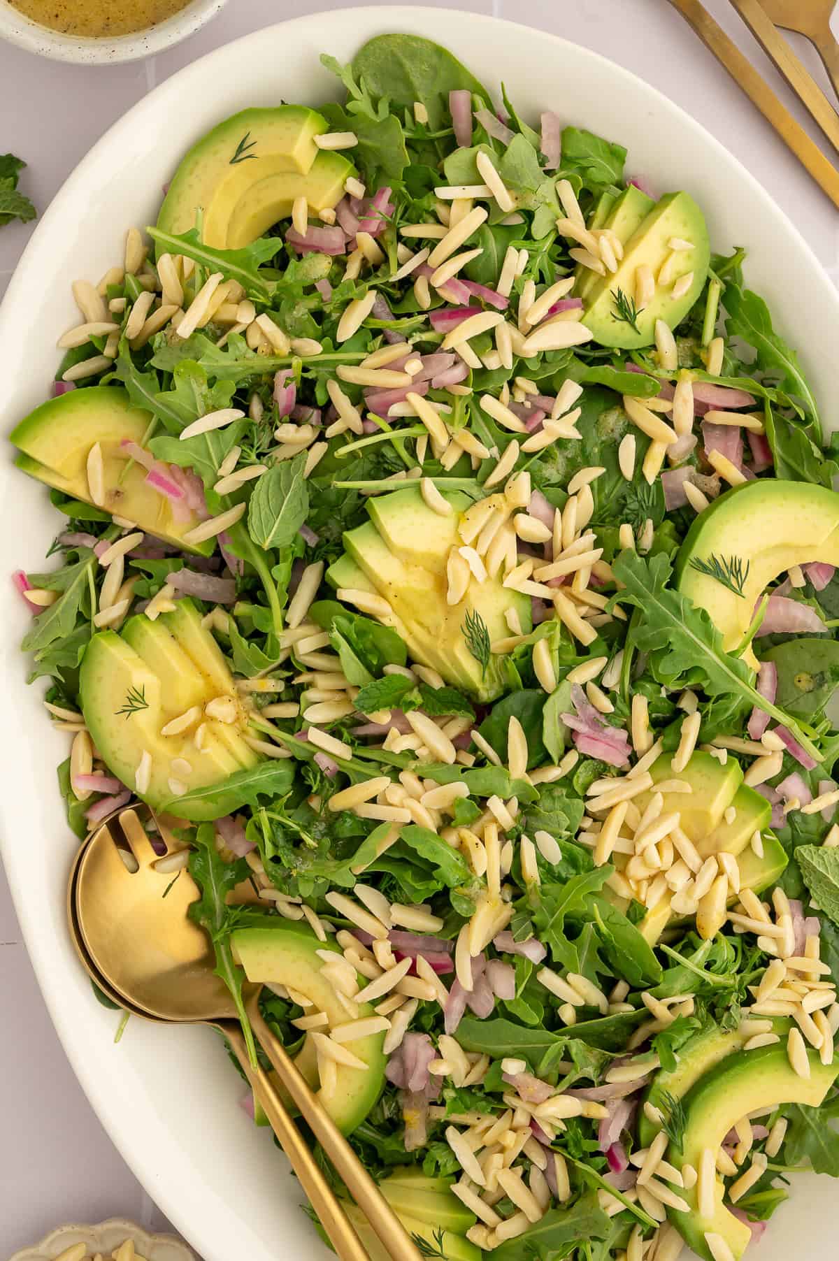 Salad on a white platter with arugula, spinach, slivered almonds, dill, mint, and avocado.