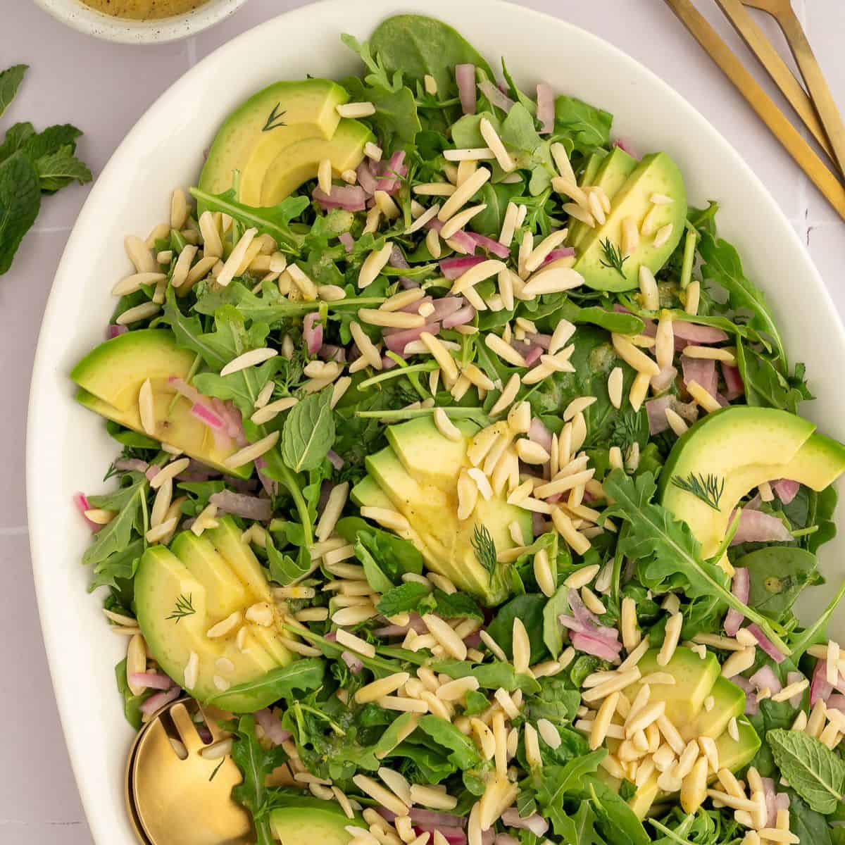 Salad on a white platter with arugula, spinach, slivered almonds, dill, mint, and avocado.