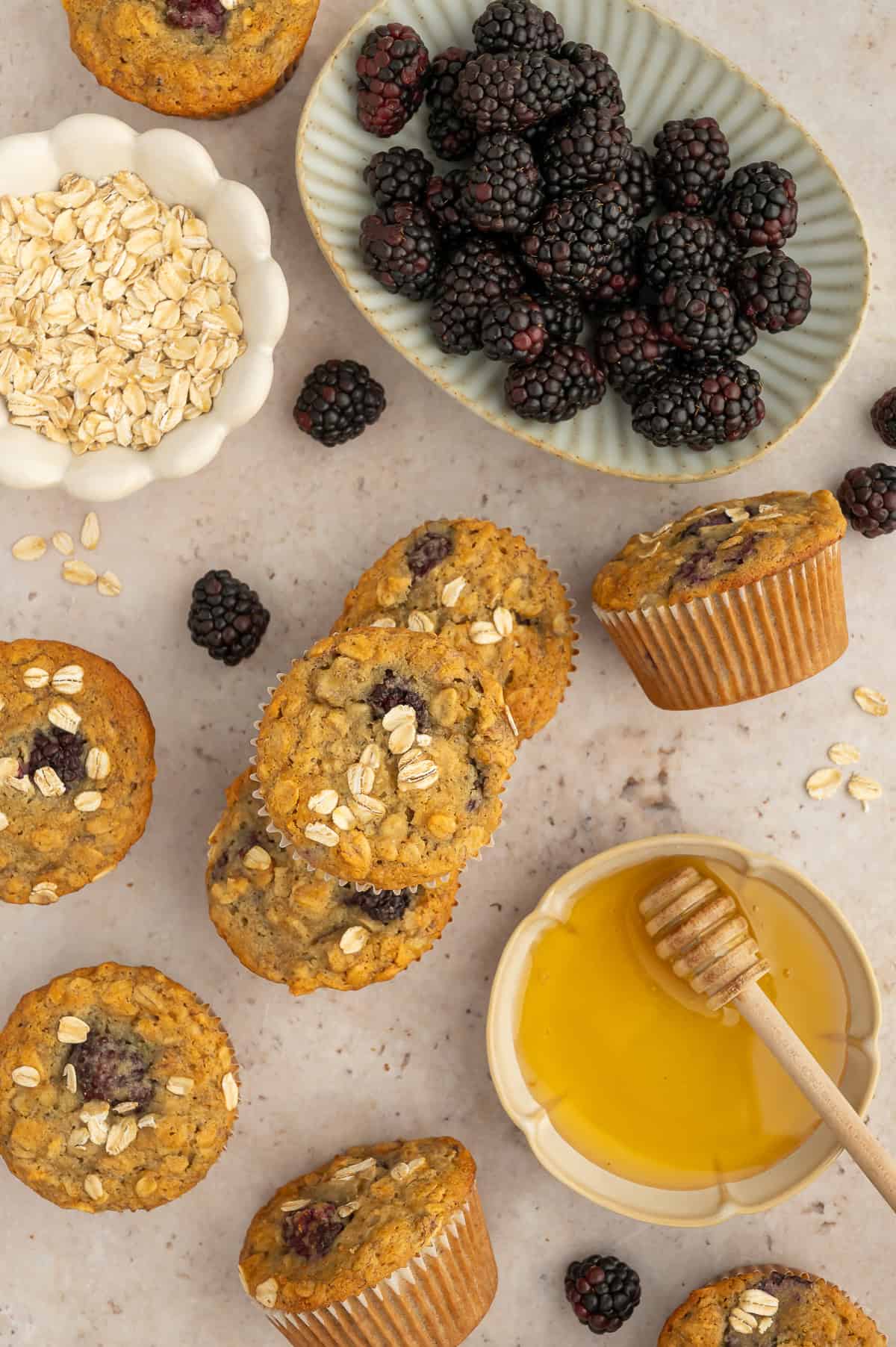 Top down view of banana blackberry oatmeal muffins with oats on top, next to bowls of money, oats, and blackberries.