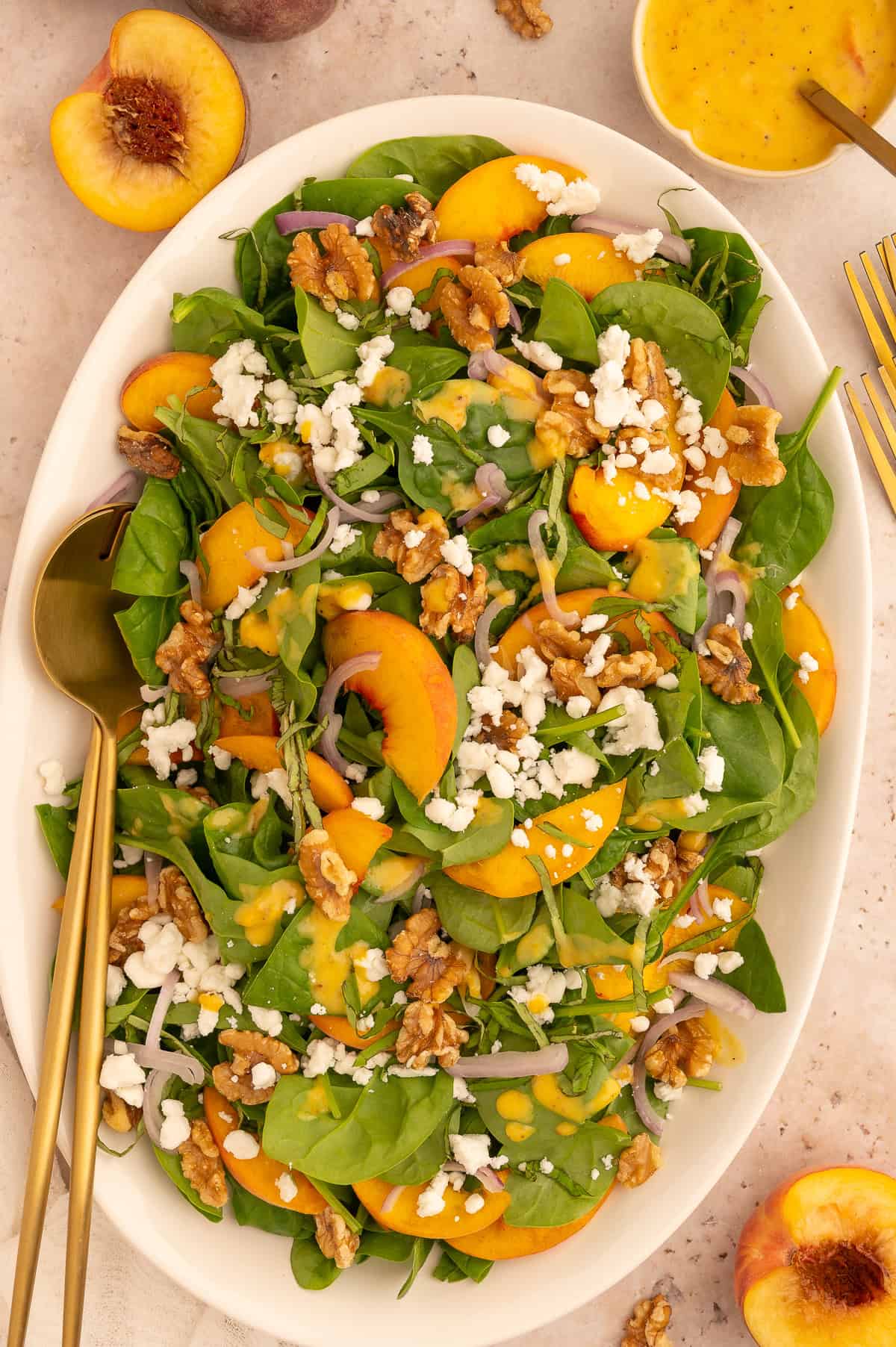 Colorful peach and walnut salad on a large platter topped with goat cheese with gold serving spoons.