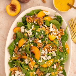 Colorful peach and walnut salad on a large platter topped with goat cheese, next to gold forks and sliced peaches.