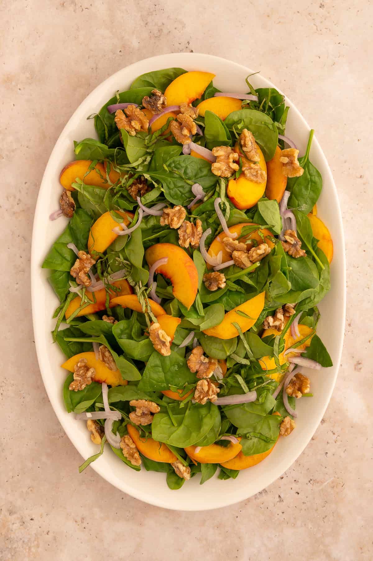 Toasted walnuts and sliced shallot added to salad with sliced peaches, basil, and spinach on a large white platter.
