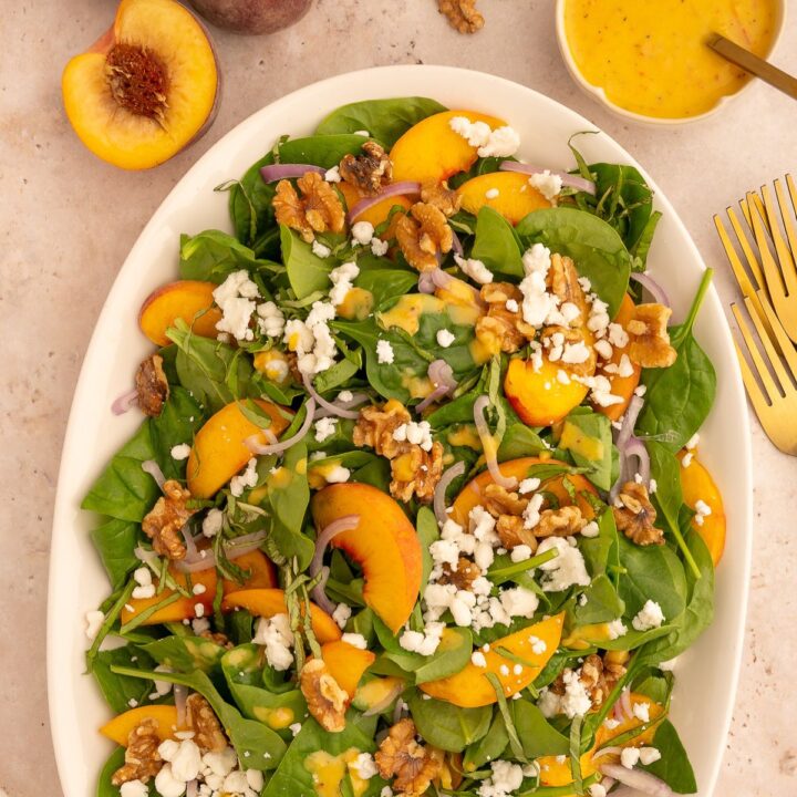 Colorful peach and walnut salad on a large platter topped with goat cheese, next to gold forks and sliced peaches.