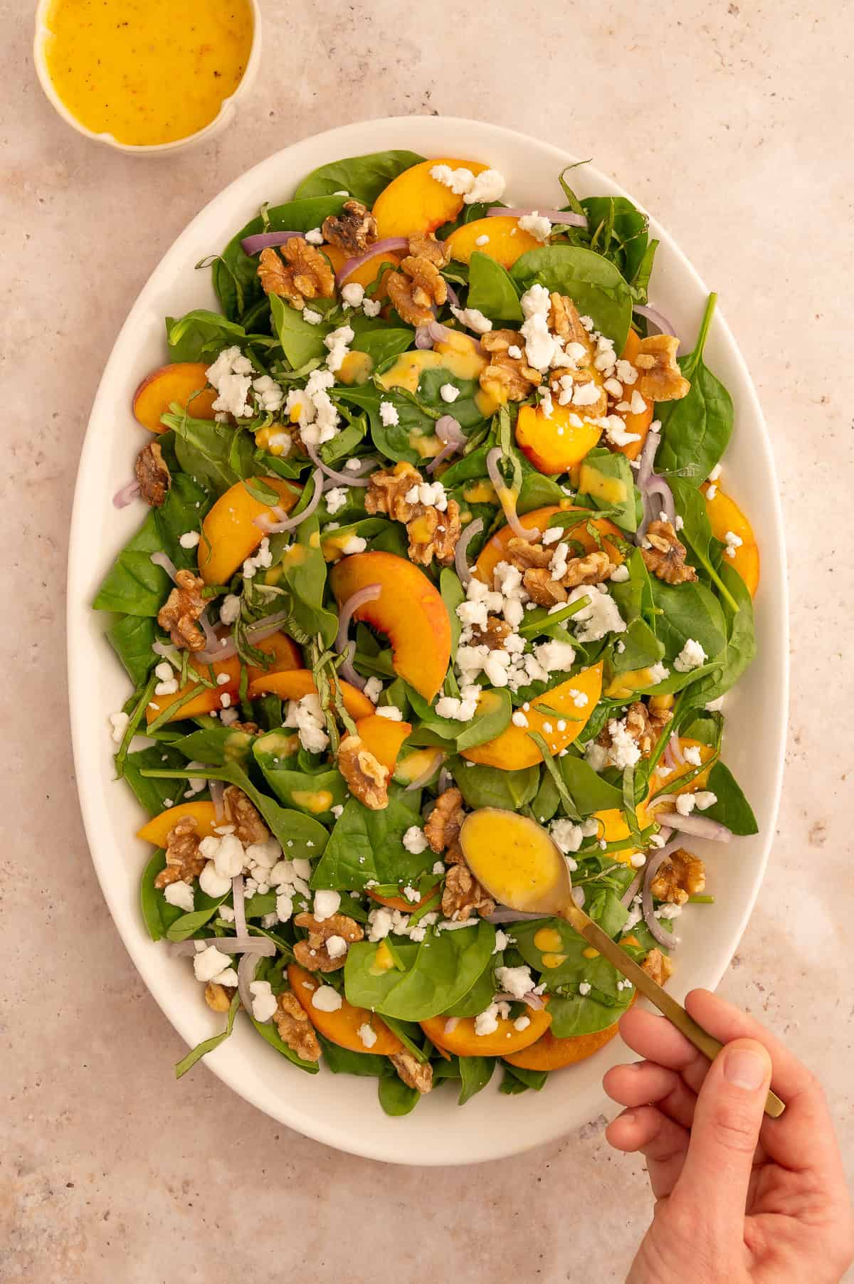 Goat cheese and peach dressing added to salad with toasted walnuts, shallots, sliced peaches, basil, and spinach on a large white platter.