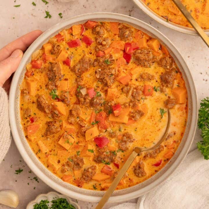 Hand holding a bowl of sweet potato and chorizo soup with a spoon in it.