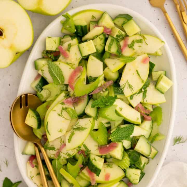 Platter of cucumber celery salad with gold serving utensils next to green apples.