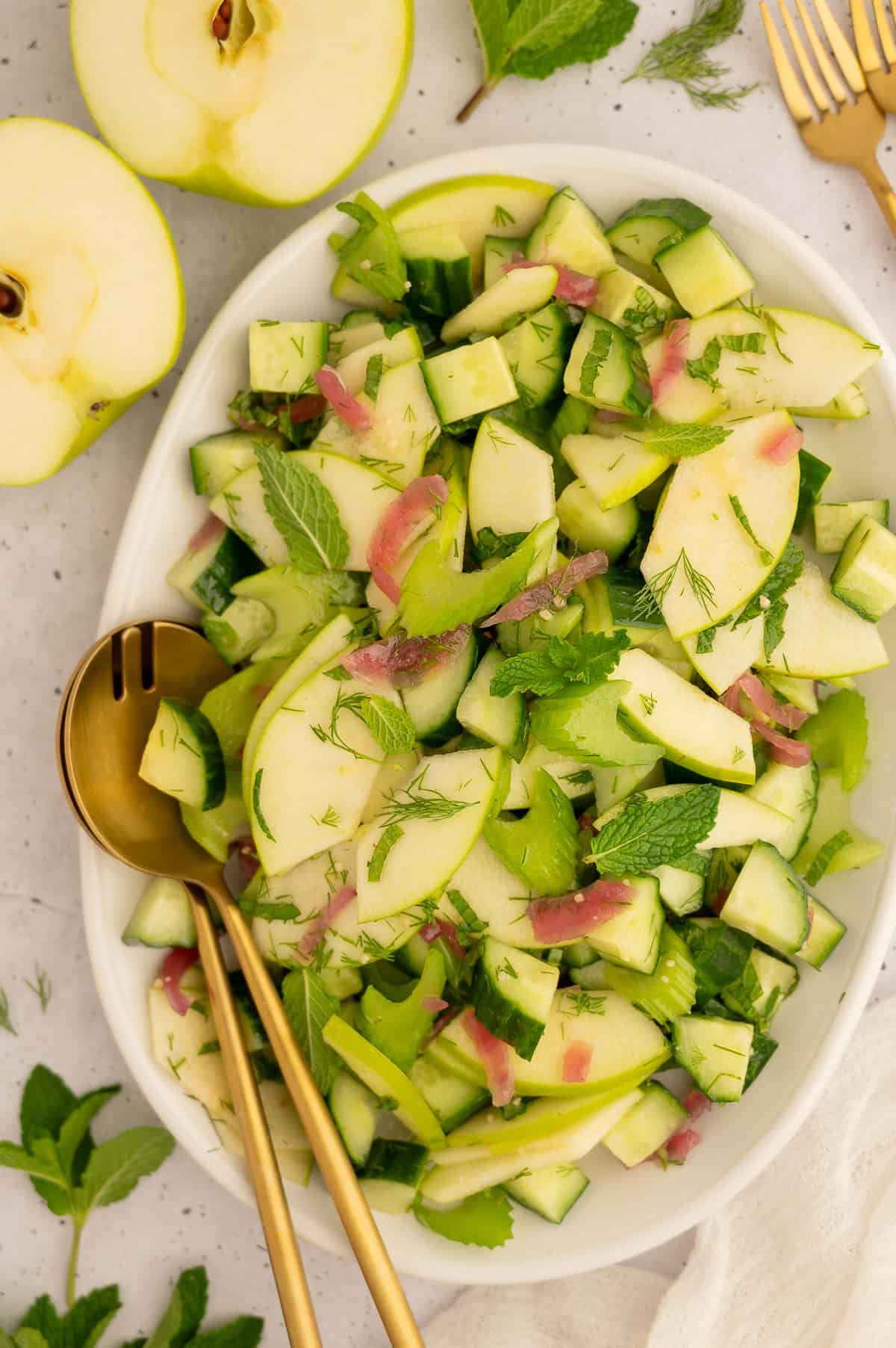 Platter of cucumber celery salad with gold serving utensils next to green apples.
